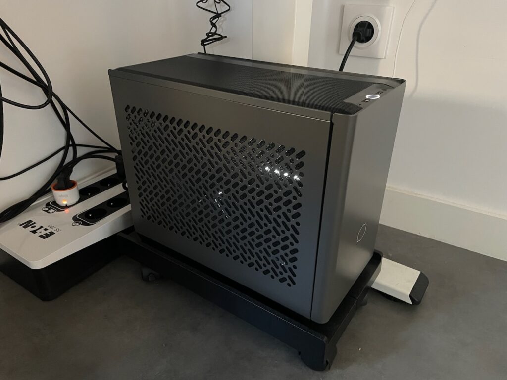 La photo montre un boîtier d'ordinateur compact Cooler Master NR200P MAX V2 positionné sur un support à roulettes noir. Il est branché sur une multiprise EI-IN 3500W, qui est elle-même connectée à une prise murale. Des câbles sont visibles à l'arrière, bien organisés. Le design perforé du panneau latéral du boîtier permet une bonne ventilation. L'ensemble est installé sur un sol en béton ciré, avec un mur blanc en arrière-plan.